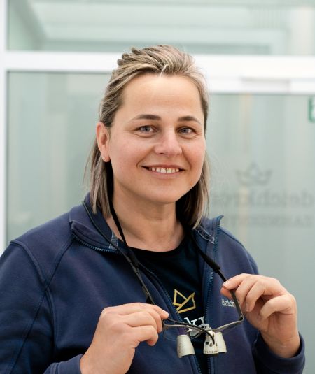Lächelnde Mitarbeiterin des Zahnzentrums Deichkrone hält Lupenbrille in der Hand