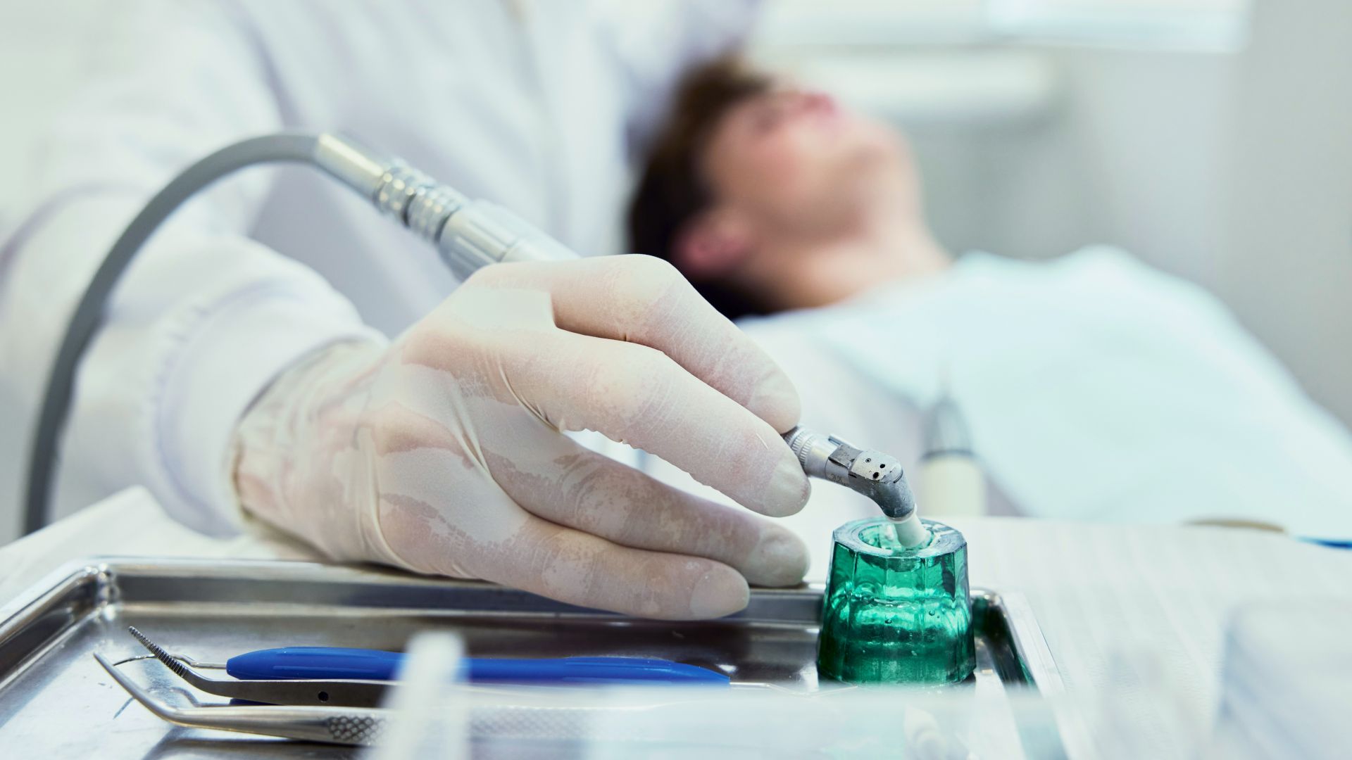 Behandelnde Hand mit Handschuh taucht zahnärztliches Instrument in Flüssigkeit mit einem Patient verschwommen im Hintergrund